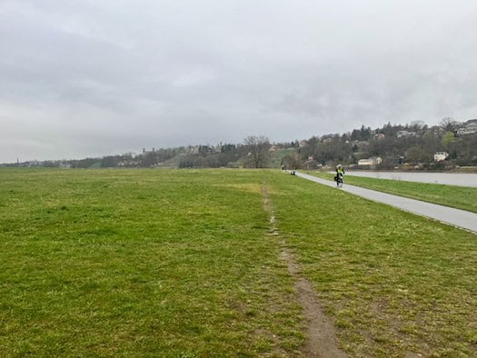 Blick auf den asphaltierten Elberadweg in Dresden bei bewölktem Himmel. Rechts fließt die Elbe, links erstrecken sich die weitläufigen Elbwiesen. Im Hintergrund sind die bewaldeten Elbhänge mit Villen und Schlössern (Albrechtsberg) zu erkennen. Ein einzelner Radfahrer ist auf dem Weg unterwegs.