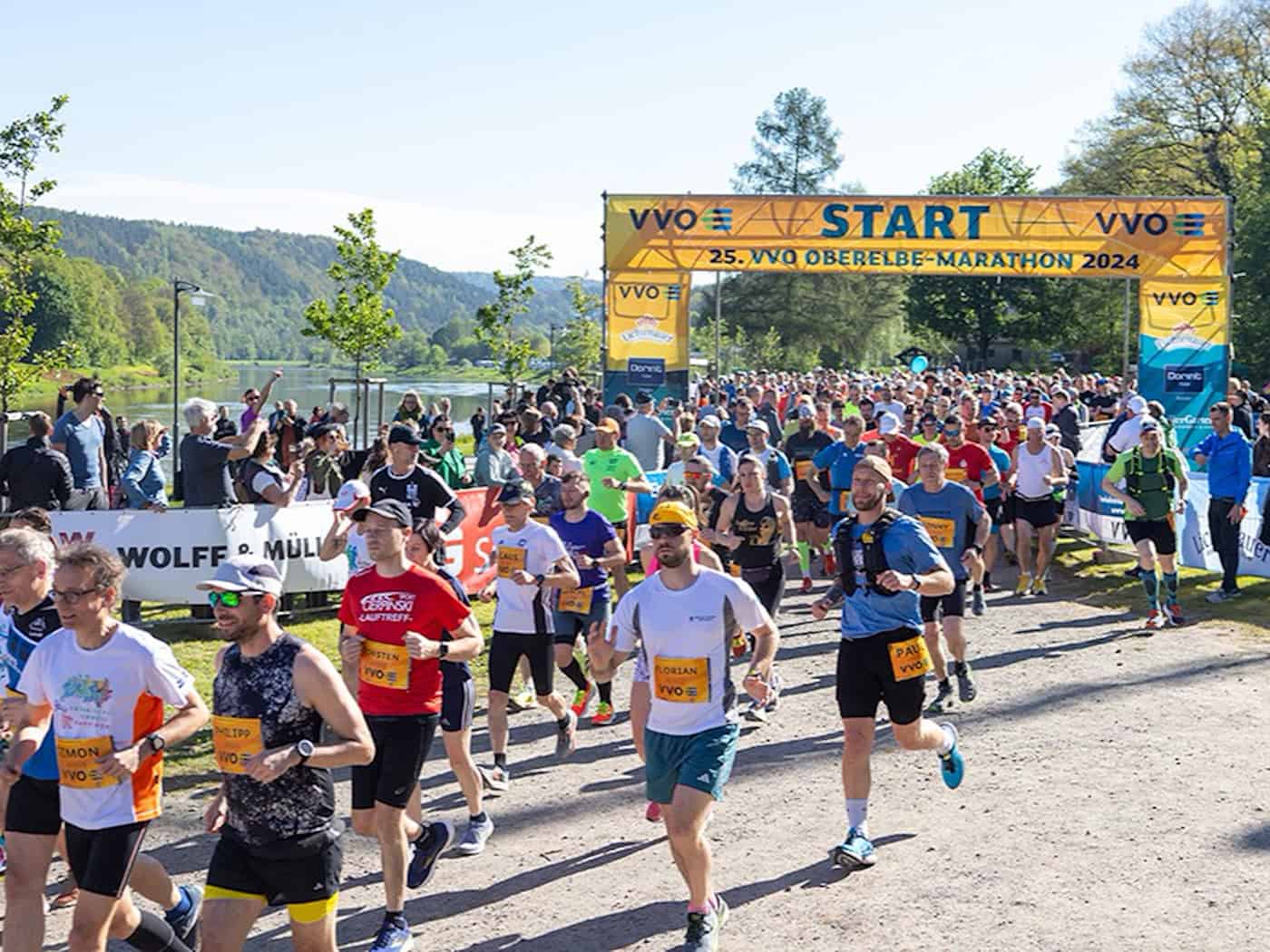 Tausende Läufer starten beim Oberelbe-Marathon unter einem großen gelben VVO-Startbogen auf einem Weg direkt an der Elbe; im Hintergrund sind bewaldete Hänge und ein strahlend blauer Himmel zu sehen.