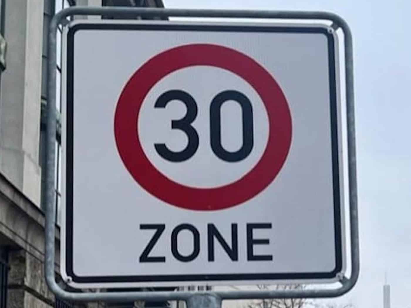 Nahaufnahme eines Verkehrsschilds für eine 30er-Zone in Dresden. Das Schild, mit einem roten Kreis um die Zahl 30 und dem Wort "ZONE" darunter, steht vor einer städtischen Kulisse mit Gebäuden und Bäumen unter grauem Himmel. Es dient als Symbol für die neue Geschwindigkeitsbegrenzung in Leubnitz-Neu