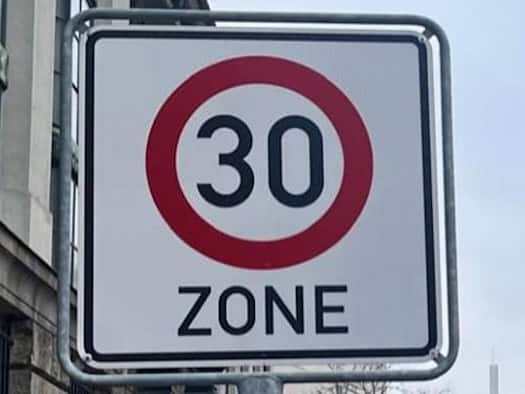Nahaufnahme eines Verkehrsschilds für eine 30er-Zone in Dresden. Das Schild, mit einem roten Kreis um die Zahl 30 und dem Wort "ZONE" darunter, steht vor einer städtischen Kulisse mit Gebäuden und Bäumen unter grauem Himmel. Es dient als Symbol für die neue Geschwindigkeitsbegrenzung in Leubnitz-Neu