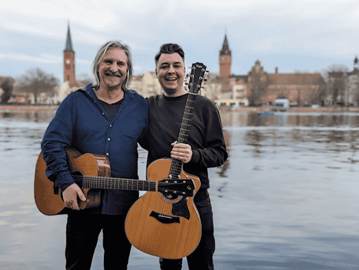 Die Musiker Dirk Zöllner und Julian Wolf lächelnd mit ihren akustischen Gitarren vor einer sommerlichen Wasser- und Stadtkulisse. Dirk Zöllner trägt ein blaues Hemd, Julian Wolf ein dunkles Sweatshirt.