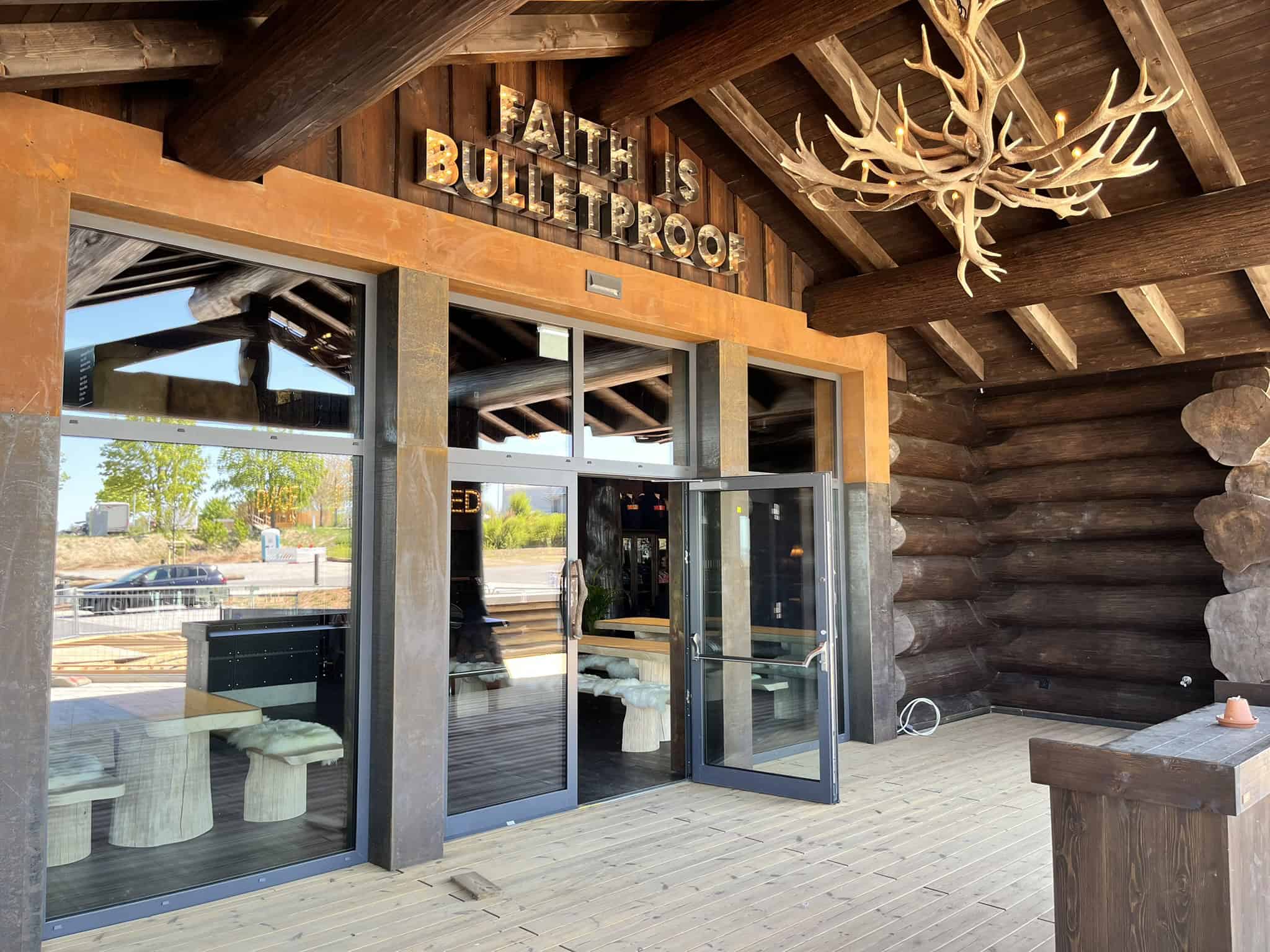 Eingangsbereich einer Blockhütte mit Glasfront und massiven Holzbalken. Über der Tür hängt der beleuchtete Schriftzug "FAITH IS BULLETPROOF". Ein großer Geweih-Kronleuchter hängt unter dem Dachvorsprung.
