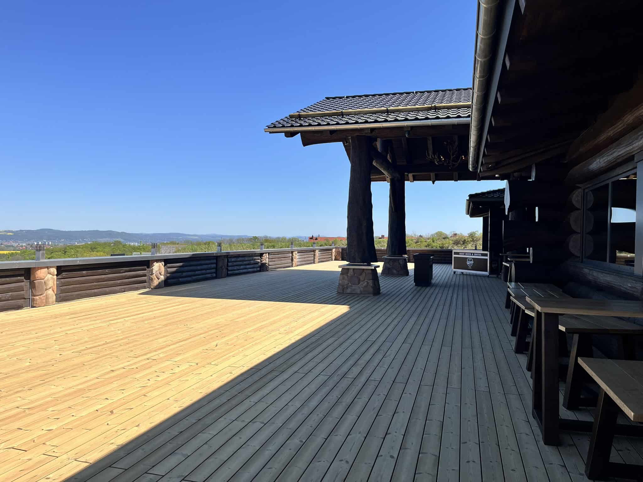 Eine weitläufige, leere Holzterrasse an einem dunklen Blockhaus-Gebäude. Massive Holzpfeiler stützen das Dach, im Hintergrund ist eine bewaldete Hügelkette unter strahlend blauem Himmel zu sehen.