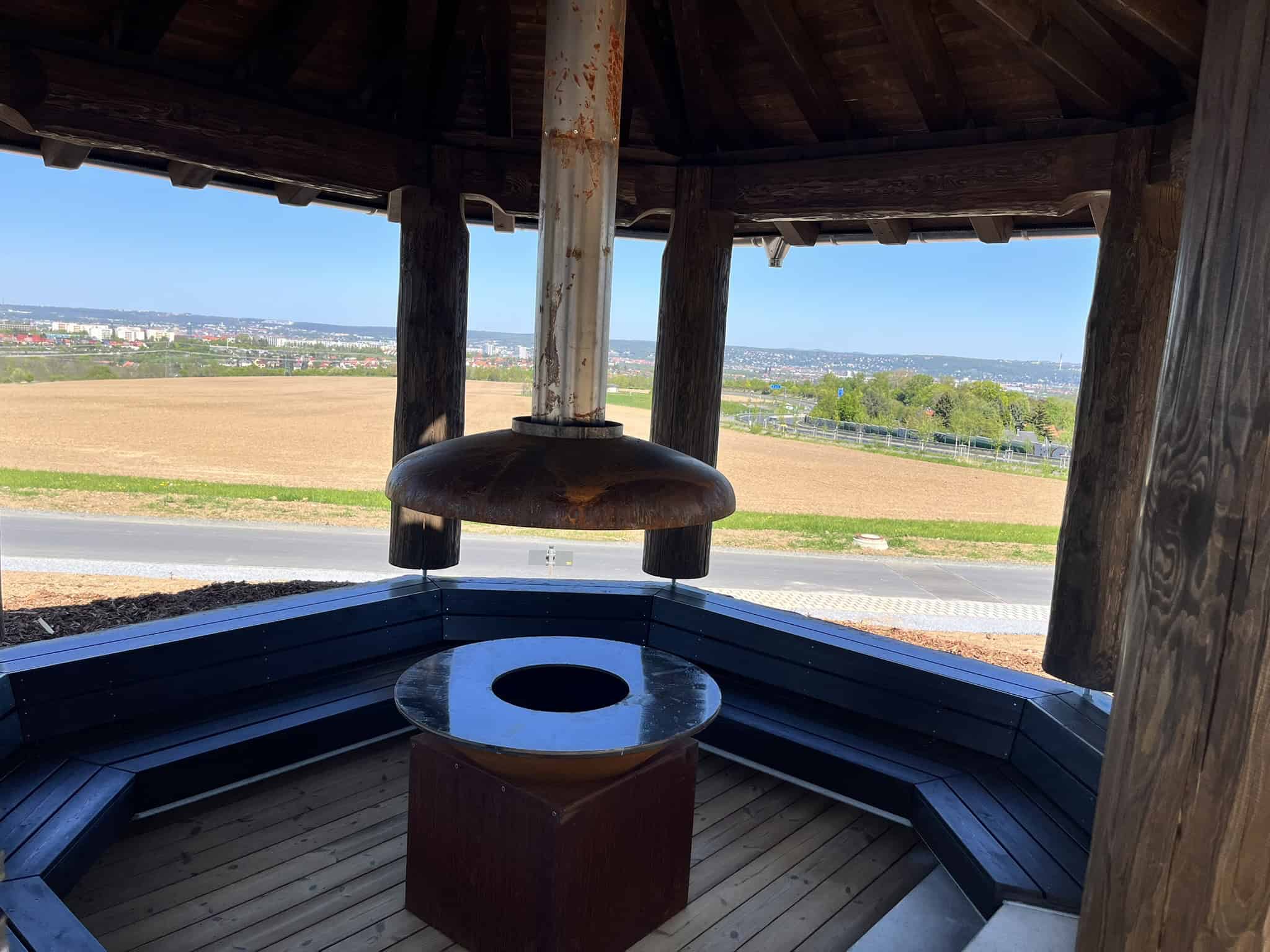 Innenansicht eines hölzernen Pavillons mit einer zentralen, modernen Feuerstelle und Abzug. Im Hintergrund erstreckt sich eine weite Landschaft mit Feldern und der Stadt Dresden unter blauem Himmel.