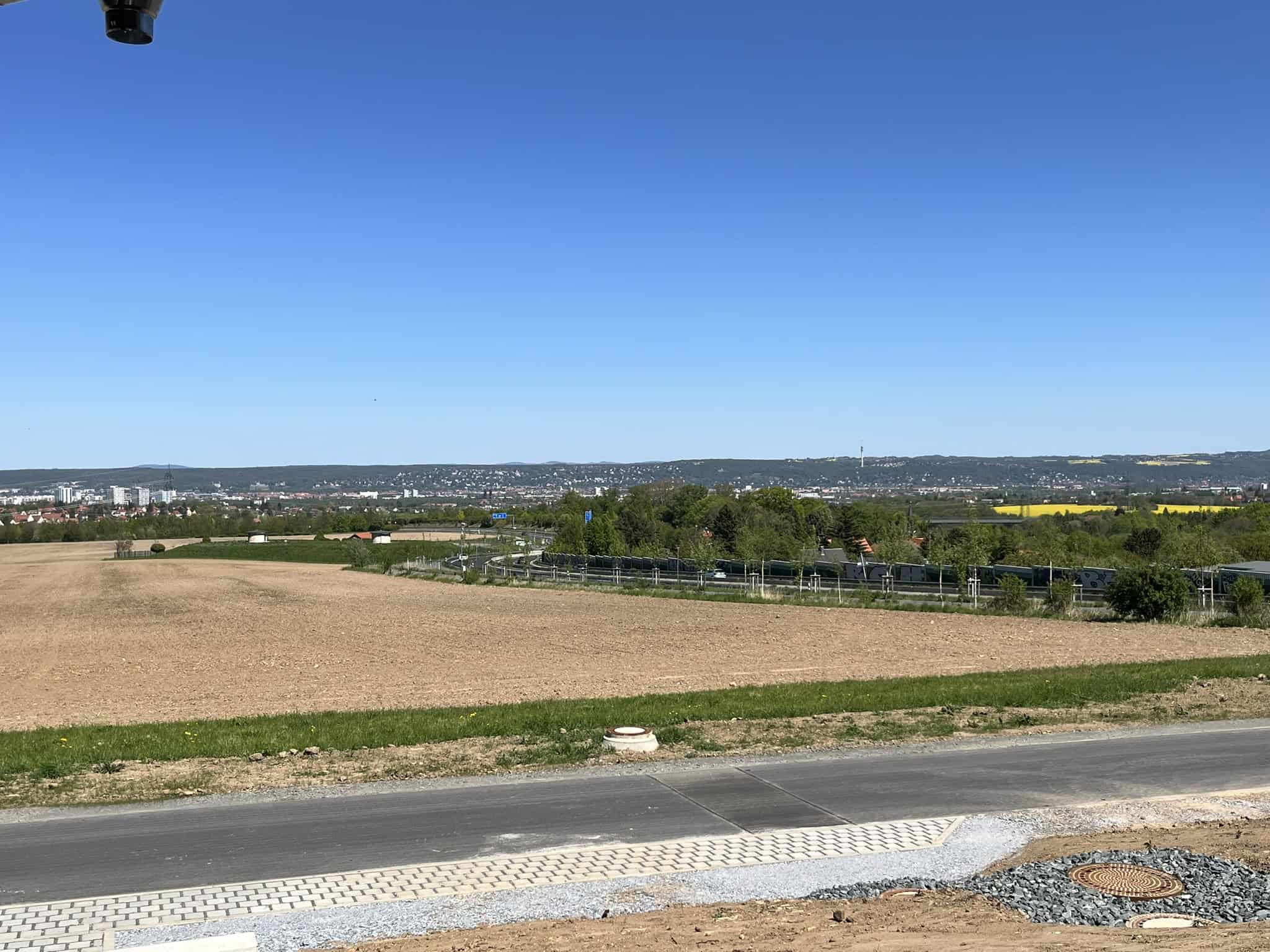 Weitwinkelaufnahme von der Terrasse des Timberjacks in Bannewitz über eine braune Ackerfläche hinweg auf die im Dunst liegende Stadt Dresden unter strahlend blauem Himmel.