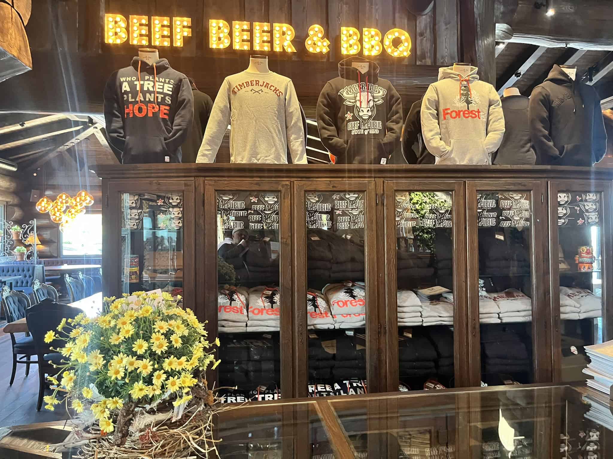 Innenansicht eines rustikalen Verkaufsbereichs mit Holzschränken, in denen Kleidung gestapelt ist. Darüber stehen Mannequins mit grauen und schwarzen Hoodies unter einem leuchtenden "BEEF BEER & BBQ" Schild.