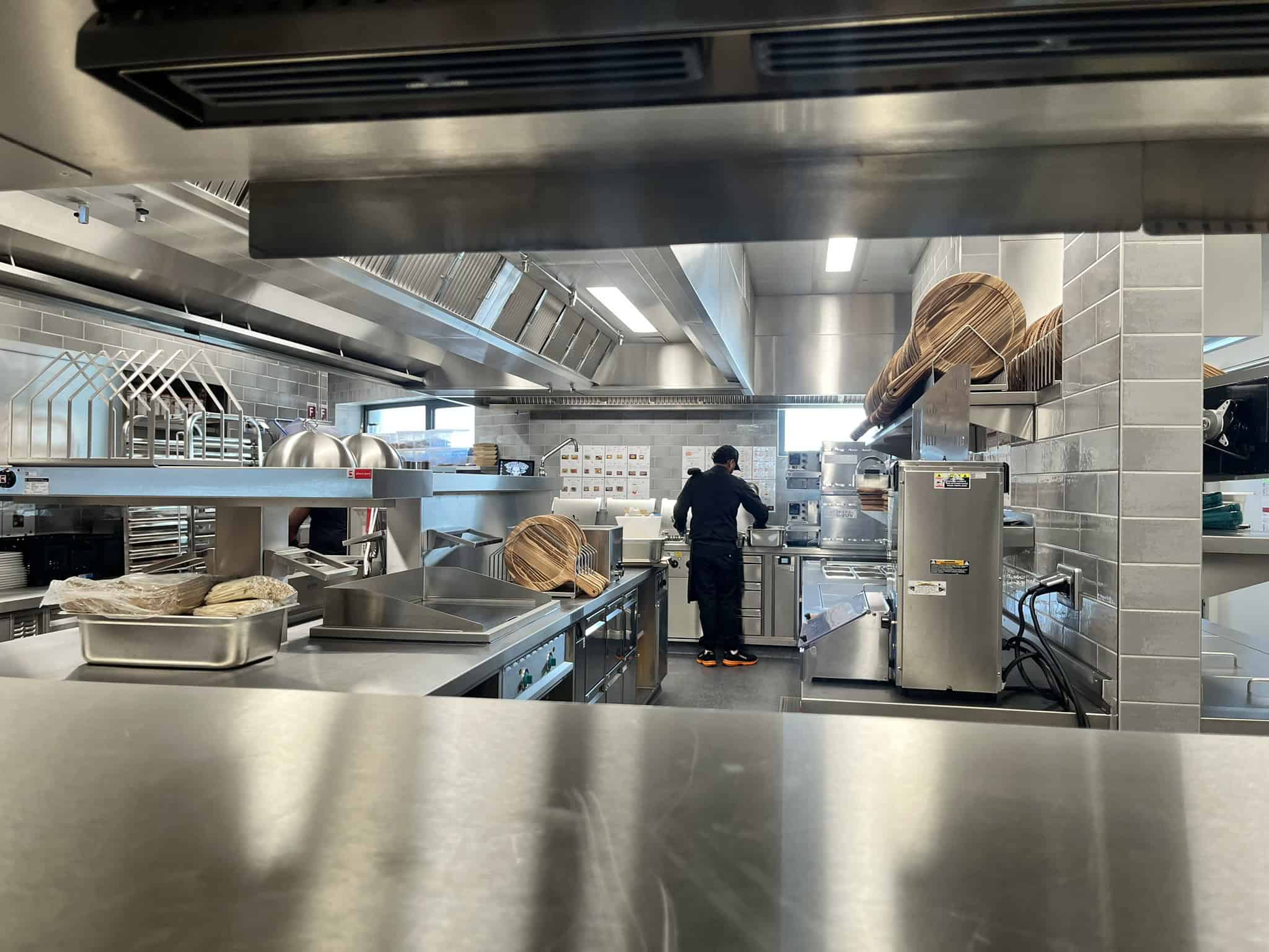 Blick in eine professionelle, saubere Gastronomie-Küche aus Edelstahl. Ein Mitarbeiter in schwarzer Arbeitskleidung steht an einem der Herde.