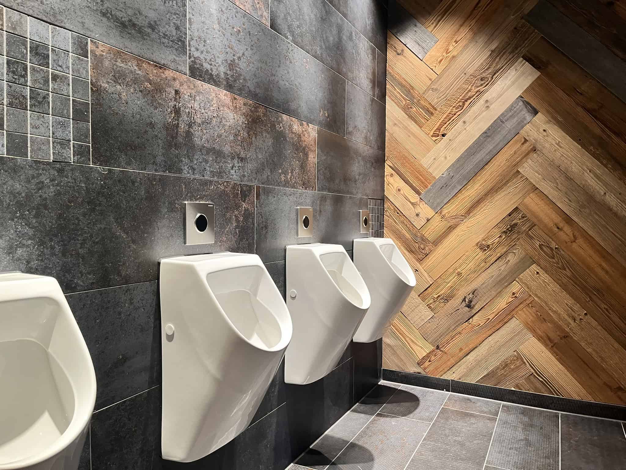 Innenansicht eines modern gestalteten Herren-WCs mit weißen Urinalen vor einer Wand aus dunklen Fliesen und Holzelementen.