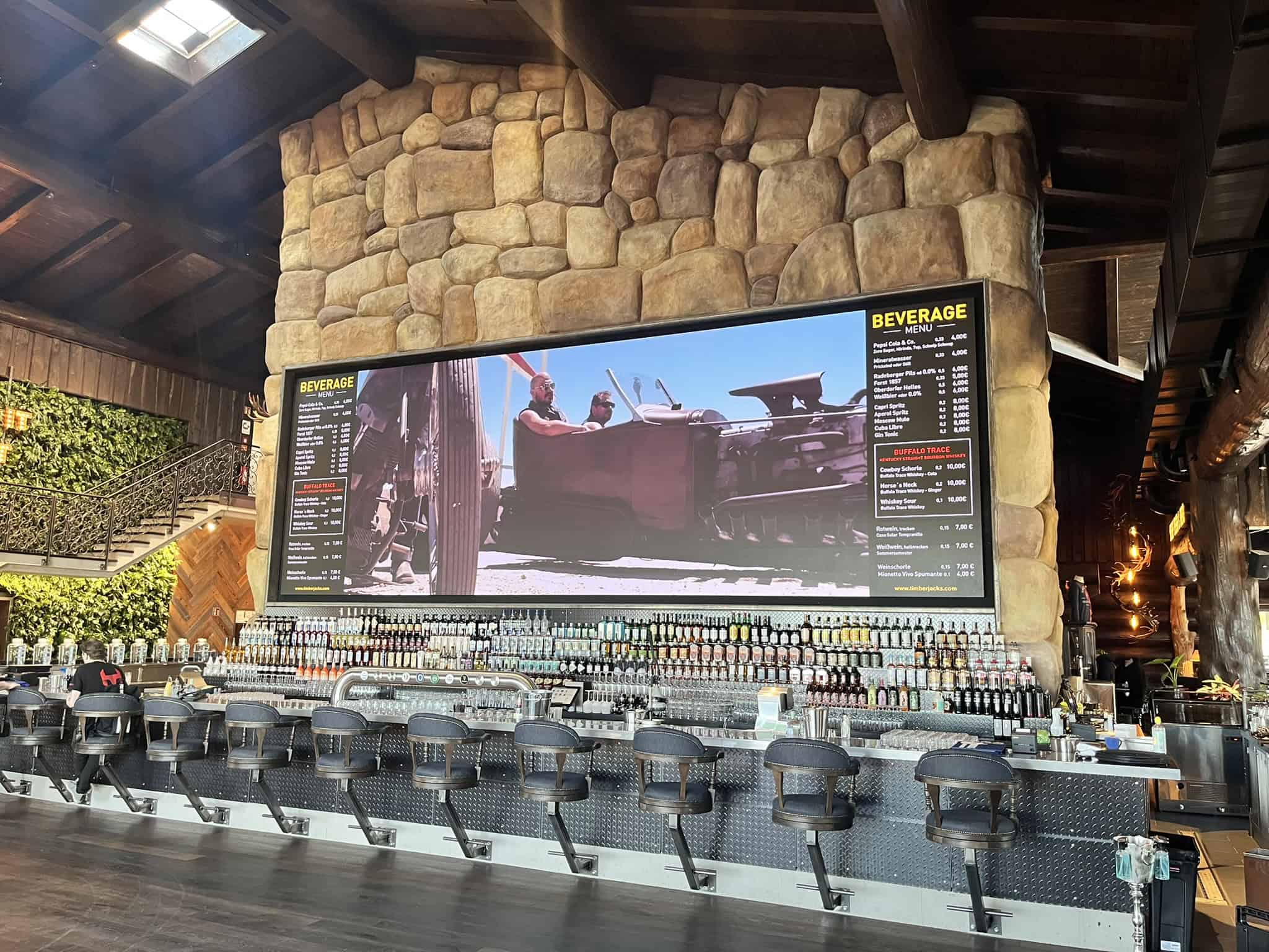 Innenansicht einer modernen Bar mit einer langen Reihe von Barhockern. Im Hintergrund eine massive Steinwand mit einer großen LED-Videowand, auf der eine Getränkekarte und ein Filmausschnitt zu sehen sind.