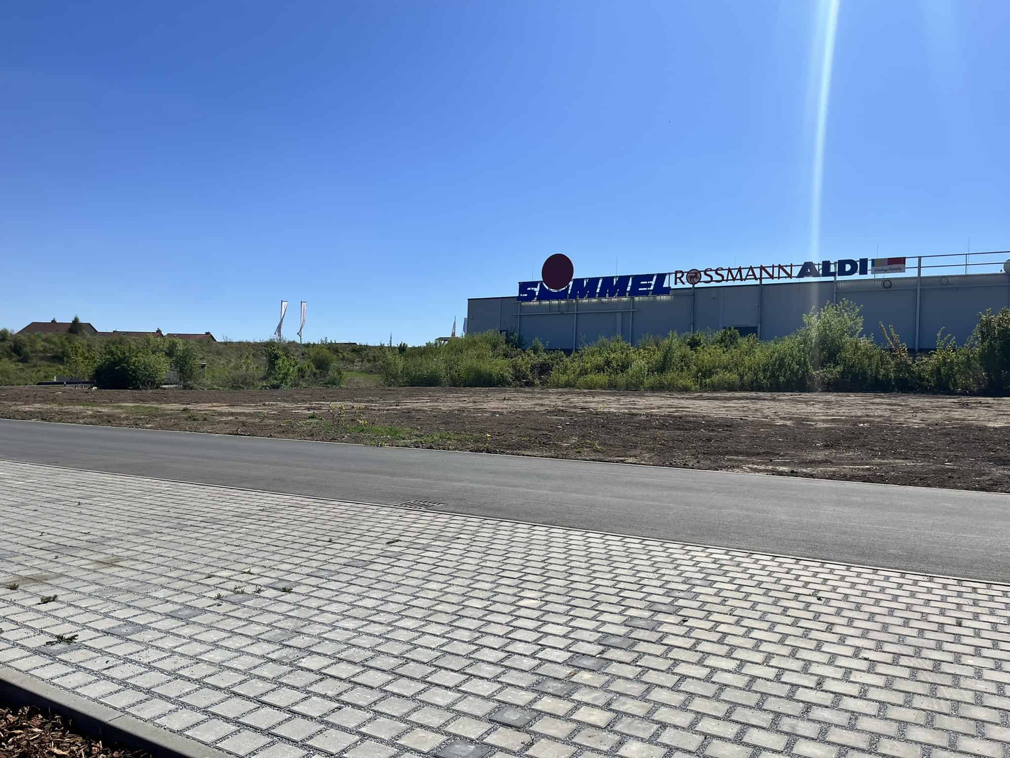 Außenansicht einer Freifläche in Bannewitz, im Hintergrund ein flacher Gewerbebau mit den Schriftzügen Simmel, Rossmann und Aldi unter blauem Himmel.