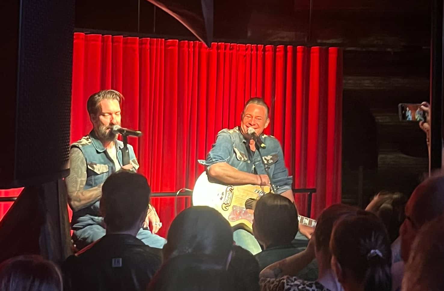 Zwei Musiker (The BossHoss) sitzen auf Barhockern auf einer Bühne vor einem roten Vorhang. Einer singt in ein Mikrofon, der andere spielt Akustikgitarre.