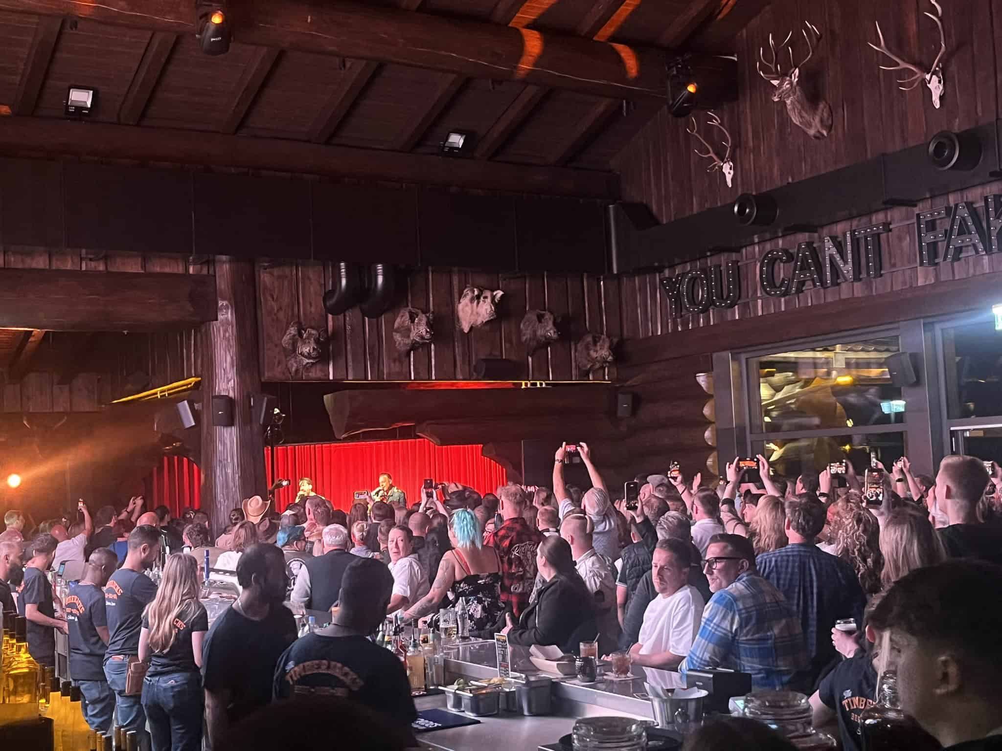 Eine Menschenmenge in einem rustikalen Blockhaus-Restaurant schaut in Richtung einer Bühne mit rotem Vorhang. Viele Gäste halten ihre Smartphones hoch, um den Auftritt zu filmen. An der Wand hängen Jagdtrophäen als Dekoration.
