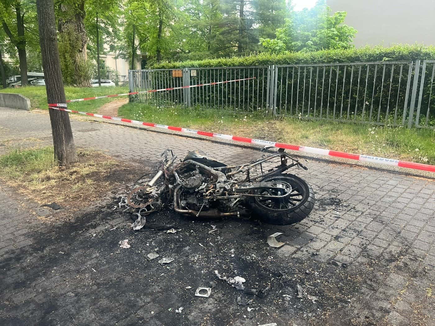 Das verkohlte Metallskelett eines ausgebrannten Motorrads liegt auf einem gepflasterten Gehweg; der Tatort ist mit rot-weißem Absperrband der Polizei gesichert.