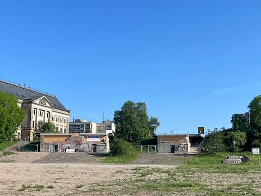 Weite, unbefestigte Fläche vor dem ehemaligen Brückenkopf der Carolabrücke in Dresden mit historischen Gebäuden im Hintergrund bei klarem Himmel.