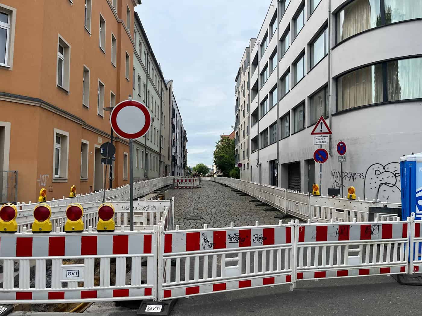 Vorbereitung Baustelle Vorwerkstraße Dresden. Blick in eine Straßenschlucht in Dresden (Vorwerkstraße) an einem bewölkten Tag. Die Straße ist beidseitig mit rot-weißen Bauzäunen und Warnleuchten für den Autoverkehr gesperrt. Auf der Straße liegt historisches Kopfsteinpflaster, im Hintergrund sind Wohnhäuser zu sehen. Ein dreieckiges Verkehrszeichen warnt vor Straßenschäden.