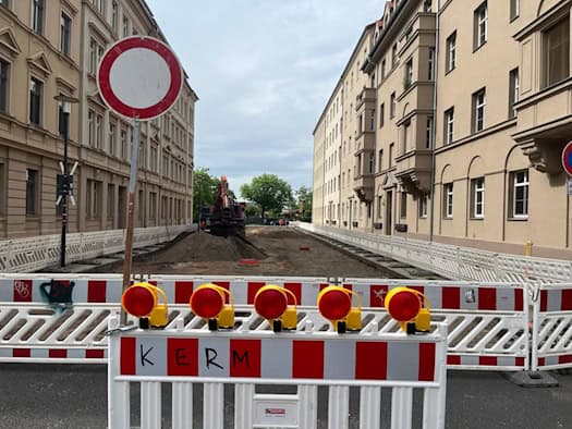 Ein Baustellenschild der Stadt Dresden steht am Straßenrand vor einem Wohngebäude. Im Hintergrund ist die abgesperrte Vorwerkstraße zu sehen, auf der Baufahrzeuge und Absperrbaken den Arbeitsbereich markieren. Ein Hinweisschild gibt Informationen zur Vollsperrung und den Einschränkungen für den Verk