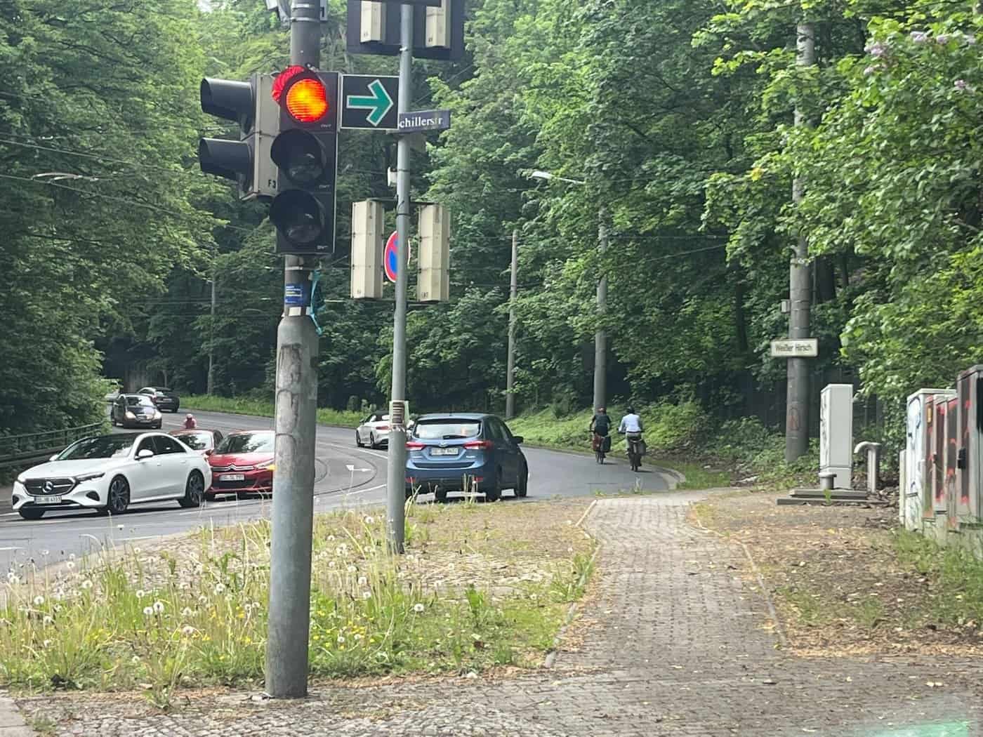 Eine Ampelanlage an der Kreuzung Bautzner Straße und Schillerstraße in Dresden. Autos und zwei Radfahrer fahren auf der asphaltierten, von dichtem Wald gesäumten Straße bergauf in Richtung Weißer Hirsch.