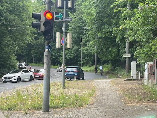 Eine Ampelanlage an der Kreuzung Bautzner Straße und Schillerstraße in Dresden. Autos und zwei Radfahrer fahren auf der asphaltierten, von dichtem Wald gesäumten Straße bergauf in Richtung Weißer Hirsch.