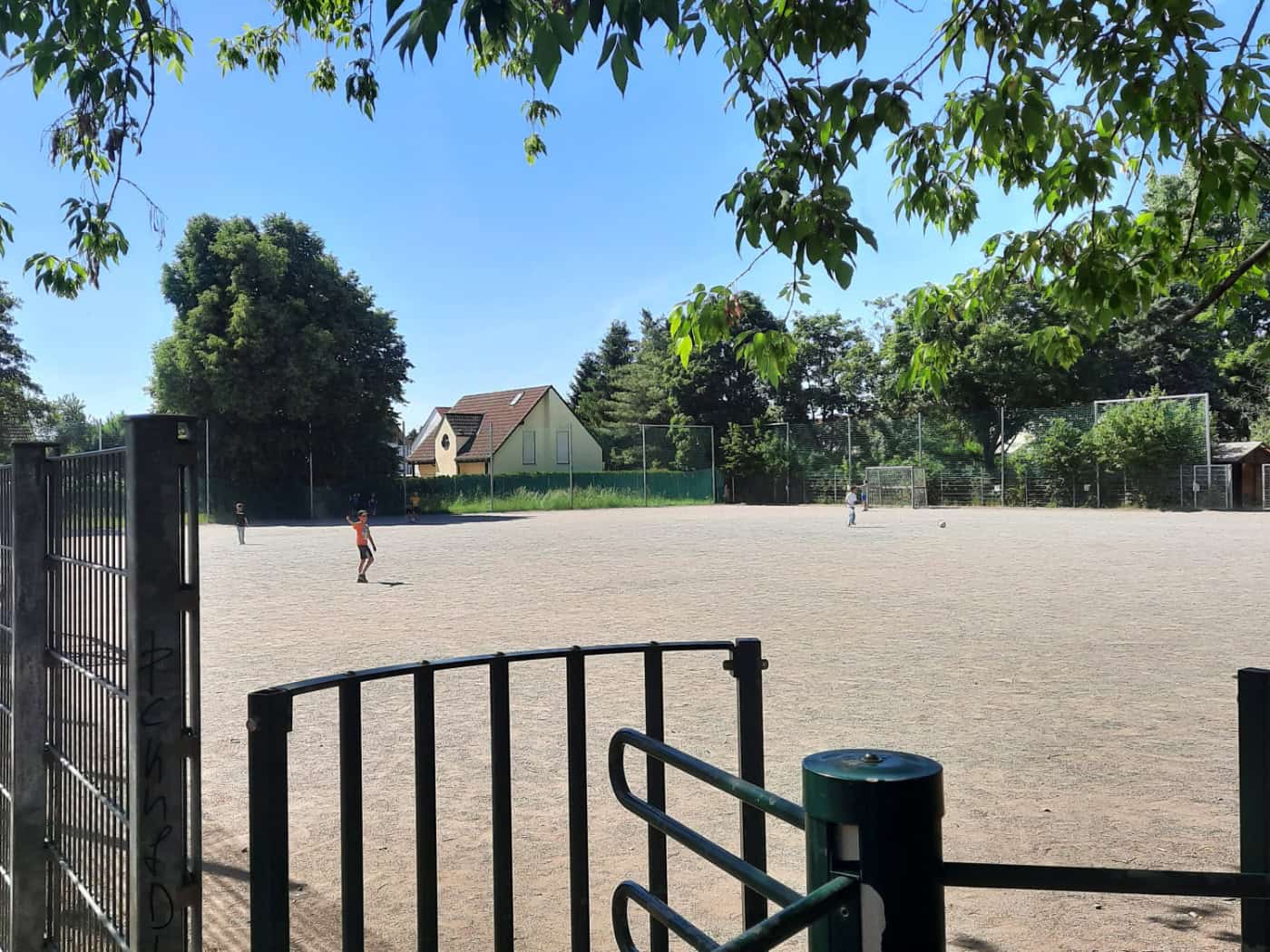 Foto: Umzäunter Fußballplatz mit Kiesboden. Grüne Bäume stehen drum herum, ein paar Kinder spielen bei blauen Himmel