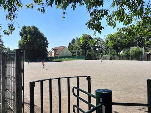 Foto: Umzäunter Fußballplatz mit Kiesboden. Grüne Bäume stehen drum herum, ein paar Kinder spielen bei blauen Himmel