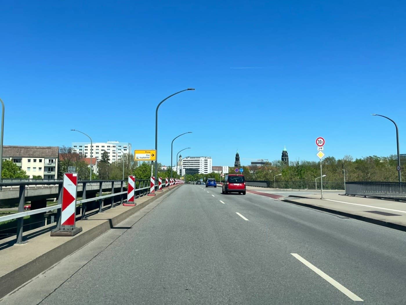 Blick über die Brücke Budapester Straße in Dresden unter wolkenlosem blauem Himmel. Rot-weiß gestreifte Warnbaken auf der linken Seite markieren die Fahrbahnverengung. Im Hintergrund sind die Silhouette der Altstadt, ein gelber Wegweiser zum Hauptbahnhof und ein roter Pkw zu sehen.