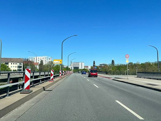 Blick über die Brücke Budapester Straße in Dresden unter wolkenlosem blauem Himmel. Rot-weiß gestreifte Warnbaken auf der linken Seite markieren die Fahrbahnverengung. Im Hintergrund sind die Silhouette der Altstadt, ein gelber Wegweiser zum Hauptbahnhof und ein roter Pkw zu sehen.