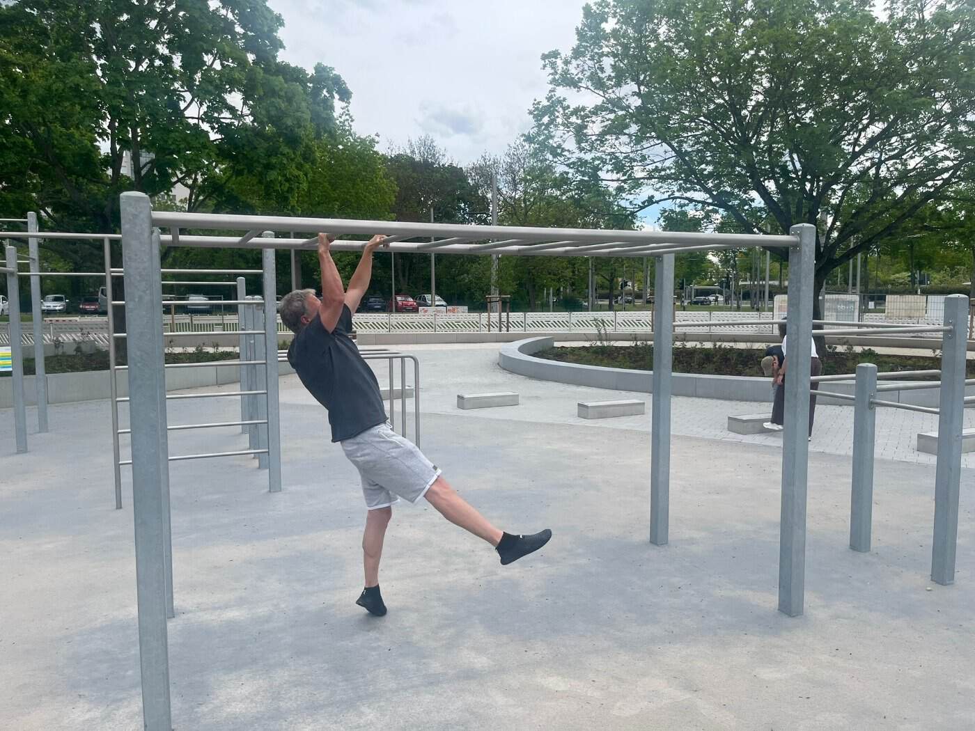 Ein Mann in kurzen grauen Hosen und schwarzem T-Shirt hängt an einer horizontalen Leiter (Monkey Bar) einer Calisthenics-Anlage im Freien und führt eine Übung aus.