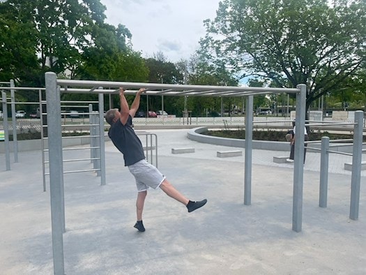 Ein Mann in kurzen grauen Hosen und schwarzem T-Shirt hängt an einer horizontalen Leiter (Monkey Bar) einer Calisthenics-Anlage im Freien und führt eine Übung aus.
