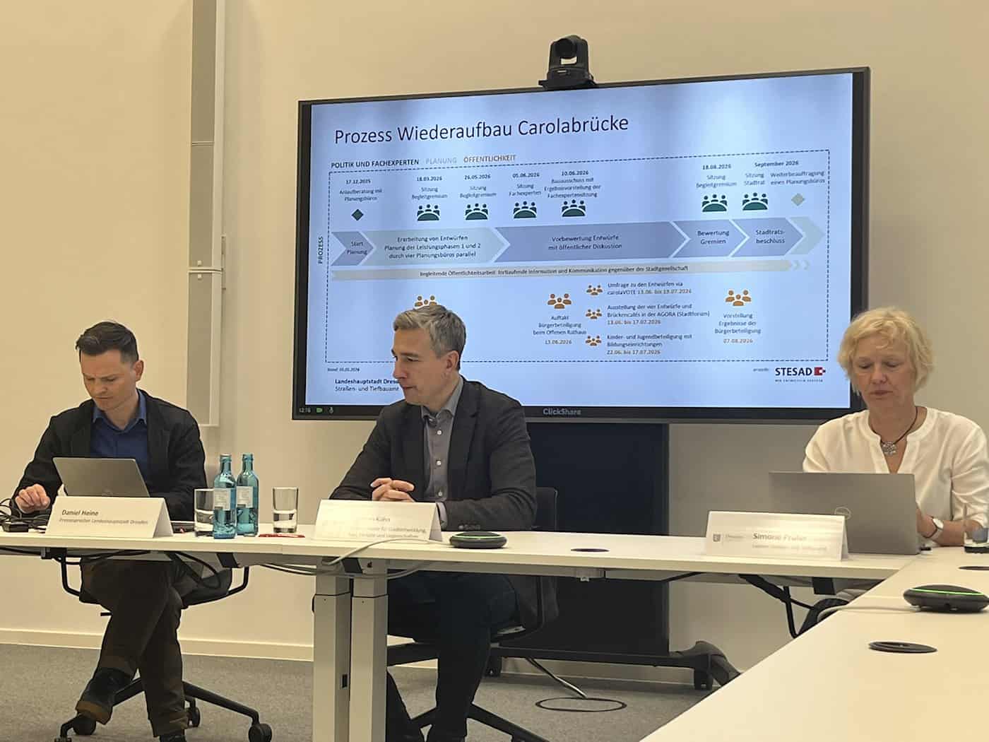 Pressesprecher Daniel Heine, Baubürgermeister Stephan Kühn und Tiefbauamtsleiterin Simone Prüfer (von links nach rechts) sitzen an einem weißen Konferenztisch. Im Hintergrund zeigt ein großer Bildschirm den grafischen Ablaufplan zum Wiederaufbau der Carolabrücke.