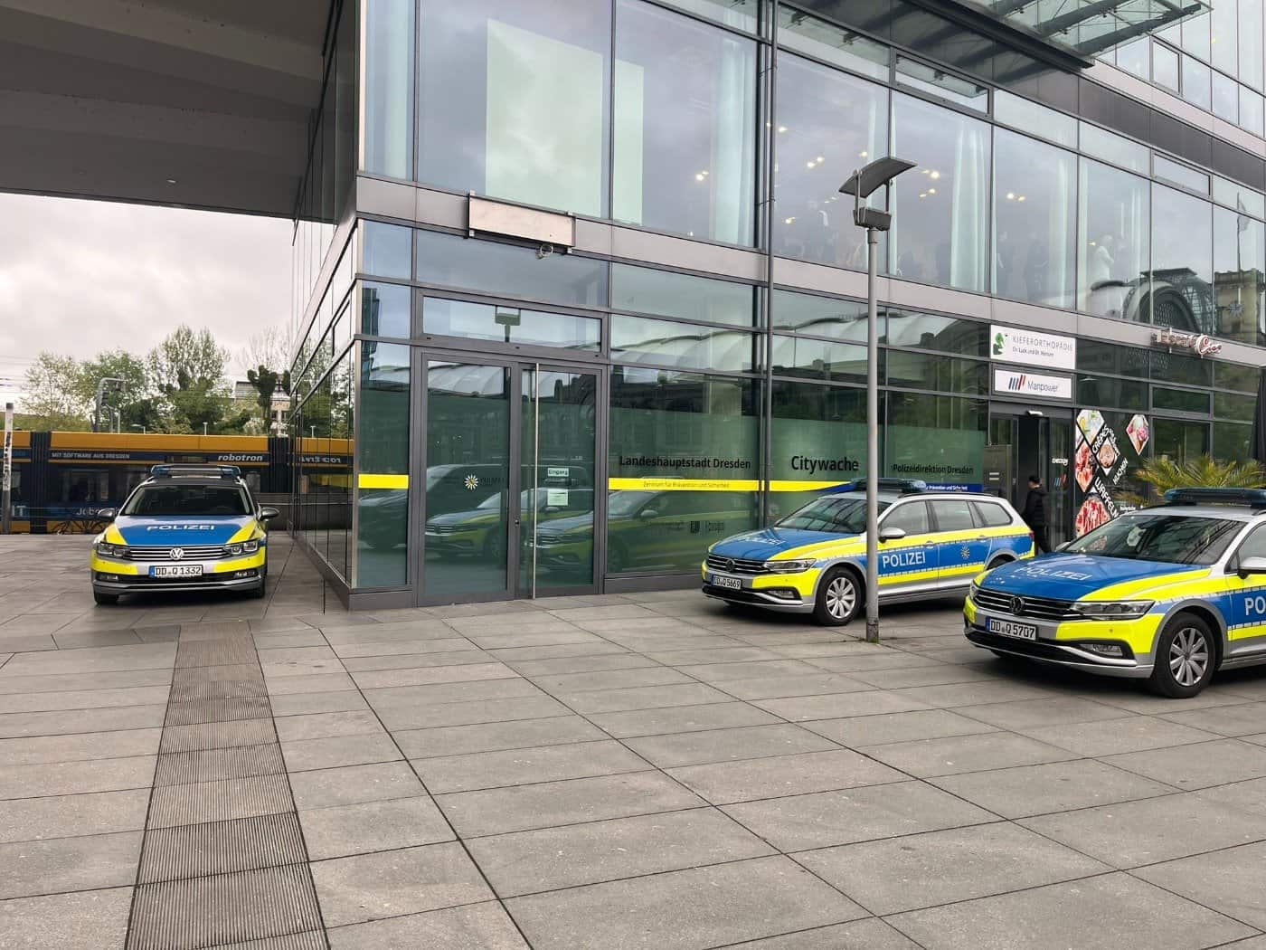 Eingang der Citywache Dresden im Erdgeschoss eines modernen Glasgebäudes (Prager Spitze). Drei blau-silber-gelbe VW Passat Polizeiautos der sächsischen Polizei sind davor geparkt. Schilder an der Tür weisen auf Landeshauptstadt Dresden und Polizeidirektion hin.