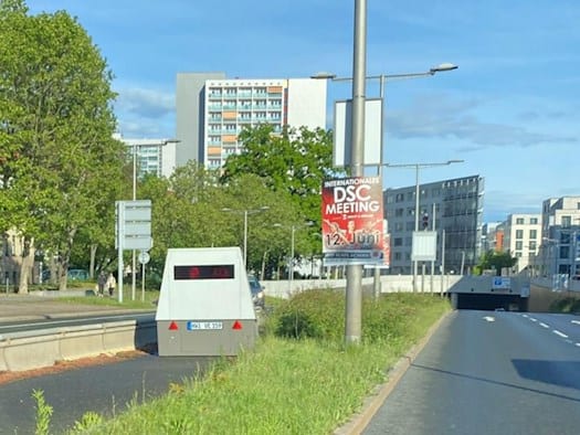 Perspektive aus einem Fahrzeug auf einen grau-weißen Blitzer-Anhänger, der auf dem Grünstreifen der Ammonstraße steht. Im Hintergrund ist ein Tunnelportal und ein Werbeplakat für ein Sport-Meeting zu sehen.