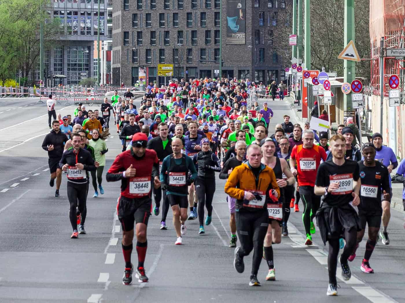 Eine große Gruppe von Marathonläufern rennt auf einer breiten Stadtstraße. Im Vordergrund sind Läufer in sportlicher Bekleidung mit Startnummern zu sehen. Im Hintergrund ragen moderne Hochhäuser und ein Hotelgebäude mit der Aufschrift „Motel One“ auf.
