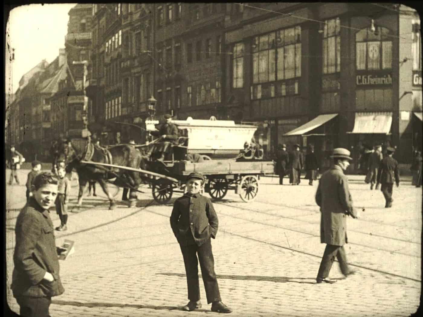 Historische Schwarz-Weiß-Aufnahme (Filmstill) von 1919: Ein offener Behelfsomnibus, besetzt mit zahlreichen Passagieren, steht auf einem belebten, gepflasterten Stadtplatz in Dresden. Im Hintergrund sind historische Fassaden, weitere Passanten und die Schienenwege der Straßenbahn zu erkennen.