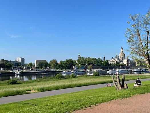 Blick über die grünen Elbwiesen und die Elbe auf die historische Silhouette der Dresdner Altstadt mit der Frauenkirche und der Kunstakademie unter wolkenlosem blauem Himmel.