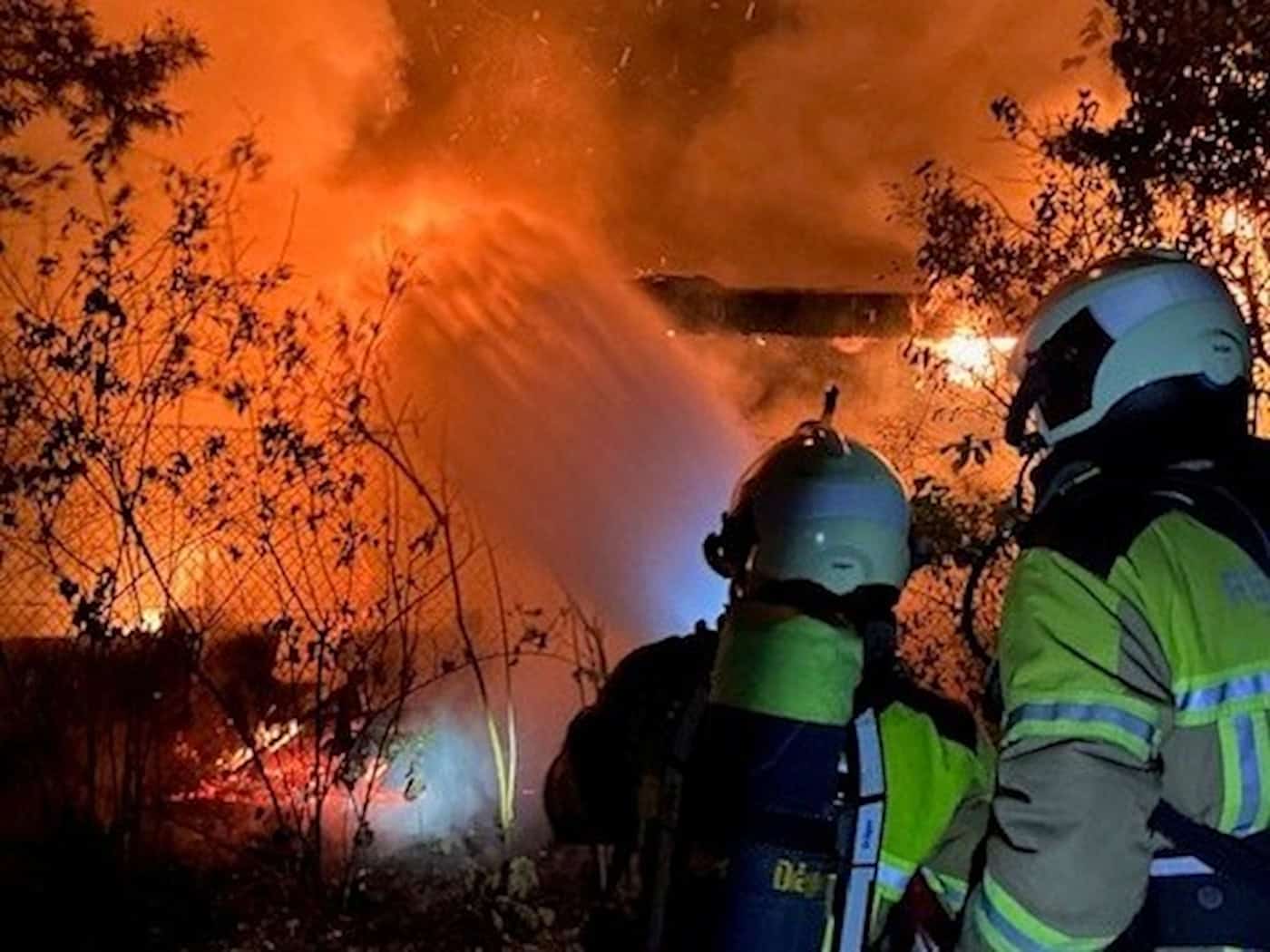 Nächtlicher Feuerwehreinsatz in einer Kleingartenanlage. Zwei Feuerwehrleute in gelb-grüner Schutzkleidung stehen im Vordergrund mit dem Rücken zum Betrachter. Einer der beiden trägt eine Atemschutzflasche auf dem Rücken. Im Hintergrund lodern helle, orangefarbene Flammen hinter Ästen und Bäumen emp