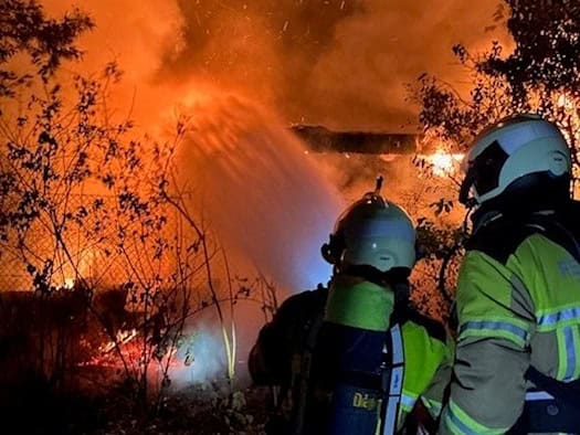 Nächtlicher Feuerwehreinsatz in einer Kleingartenanlage. Zwei Feuerwehrleute in gelb-grüner Schutzkleidung stehen im Vordergrund mit dem Rücken zum Betrachter. Einer der beiden trägt eine Atemschutzflasche auf dem Rücken. Im Hintergrund lodern helle, orangefarbene Flammen hinter Ästen und Bäumen emp