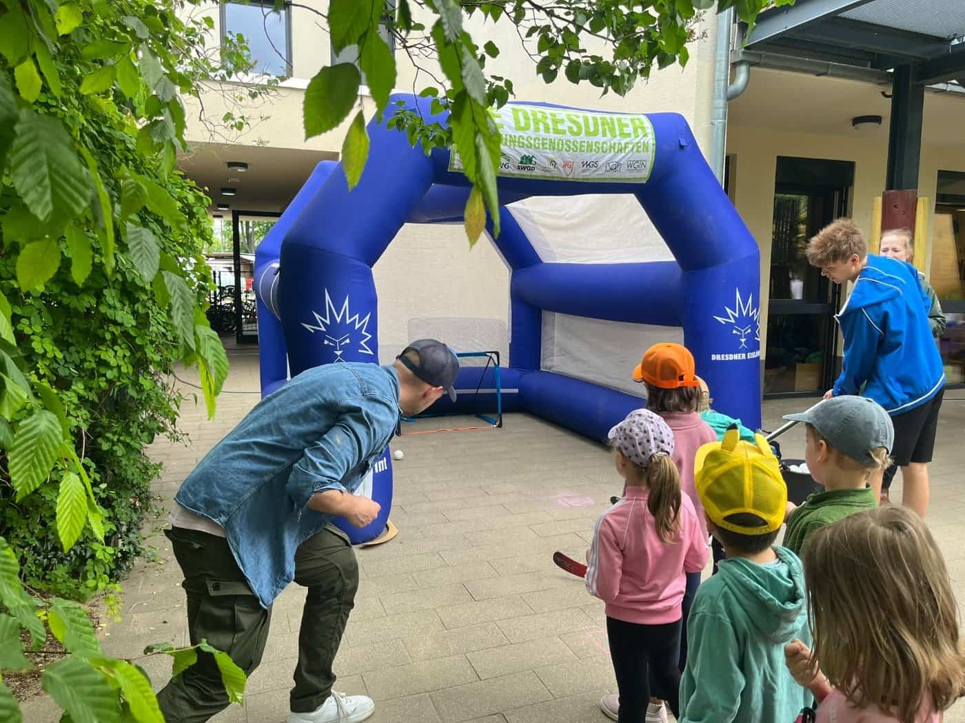 Kinder stehen mit Kappen auf dem Kopf in einer Reihe vor einem großen blauen, aufblasbaren Tor der Dresdner Eislöwen, während ein Betreuer die Station vorbereitet.