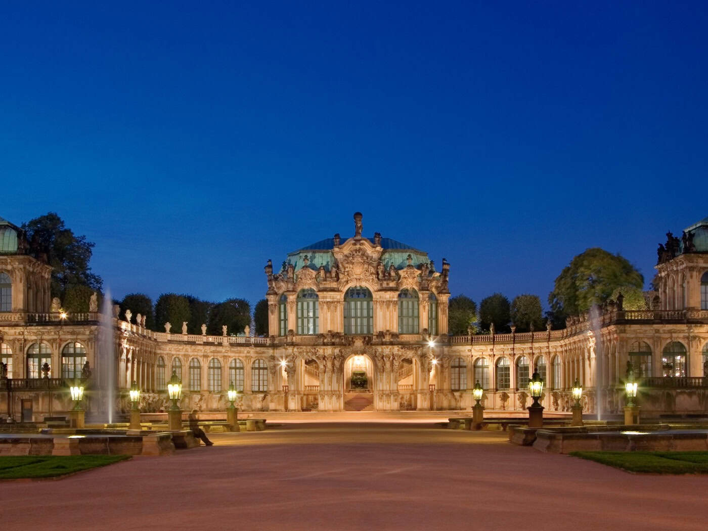 Panoramaaufnahme des Dresdner Zwingers bei Nacht unter einem tiefblauen Abendhimmel. In der Mitte thront der festlich beleuchtete Wallpavillon, rechts und links erstrecken sich die Bogengalerien. Im weitläufigen Innenhof sind zwei große, hell angestrahlte Wasserfontänen zu sehen.