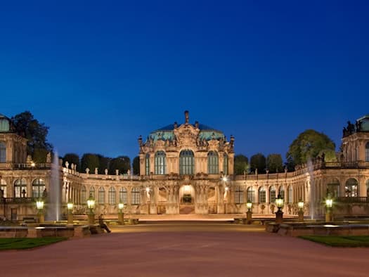 Panoramaaufnahme des Dresdner Zwingers bei Nacht unter einem tiefblauen Abendhimmel. In der Mitte thront der festlich beleuchtete Wallpavillon, rechts und links erstrecken sich die Bogengalerien. Im weitläufigen Innenhof sind zwei große, hell angestrahlte Wasserfontänen zu sehen.