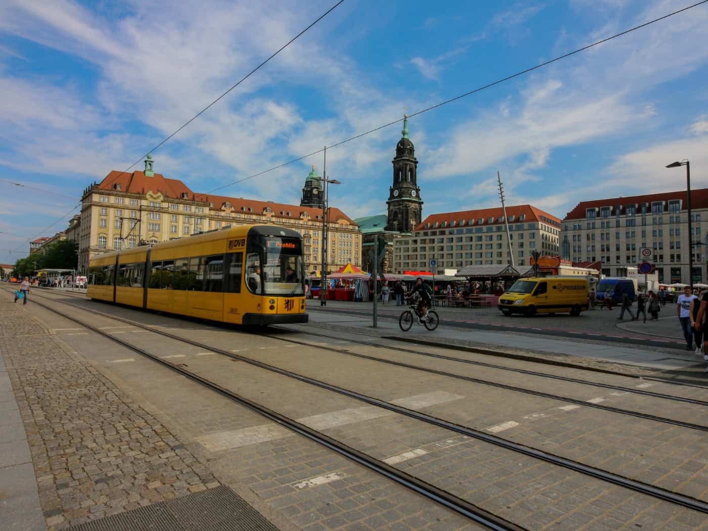 Eine gelbe Straßenbahn der DVB fährt über den Dresdner Altmarkt in Richtung Postplatz, im Hintergrund sind die Kreuzkirche und historische Gebäude unter blauem Himmel zu sehen.