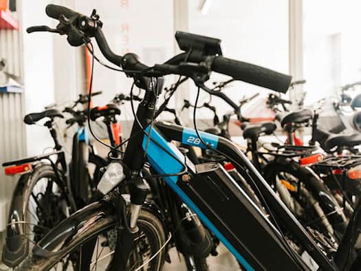 Mehrere hochwertige E-Bikes stehen dicht an dicht aufgereiht in einem Fahrradgeschäft. Im Fokus steht der Lenker und der Rahmen eines sportlichen, blau-schwarzen E-Bikes mit Akku.