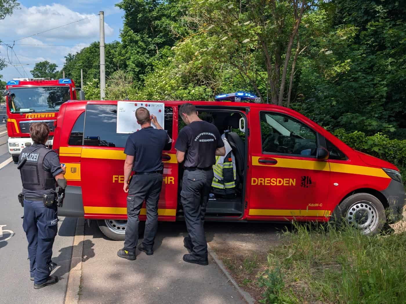 Einsatzkräfte der Feuerwehr Dresden und eine Polizeibeamtin bei der Einsatzdokumentation an einem Einsatzleitwagen (KdoW-C) während eines Brandereignisses.