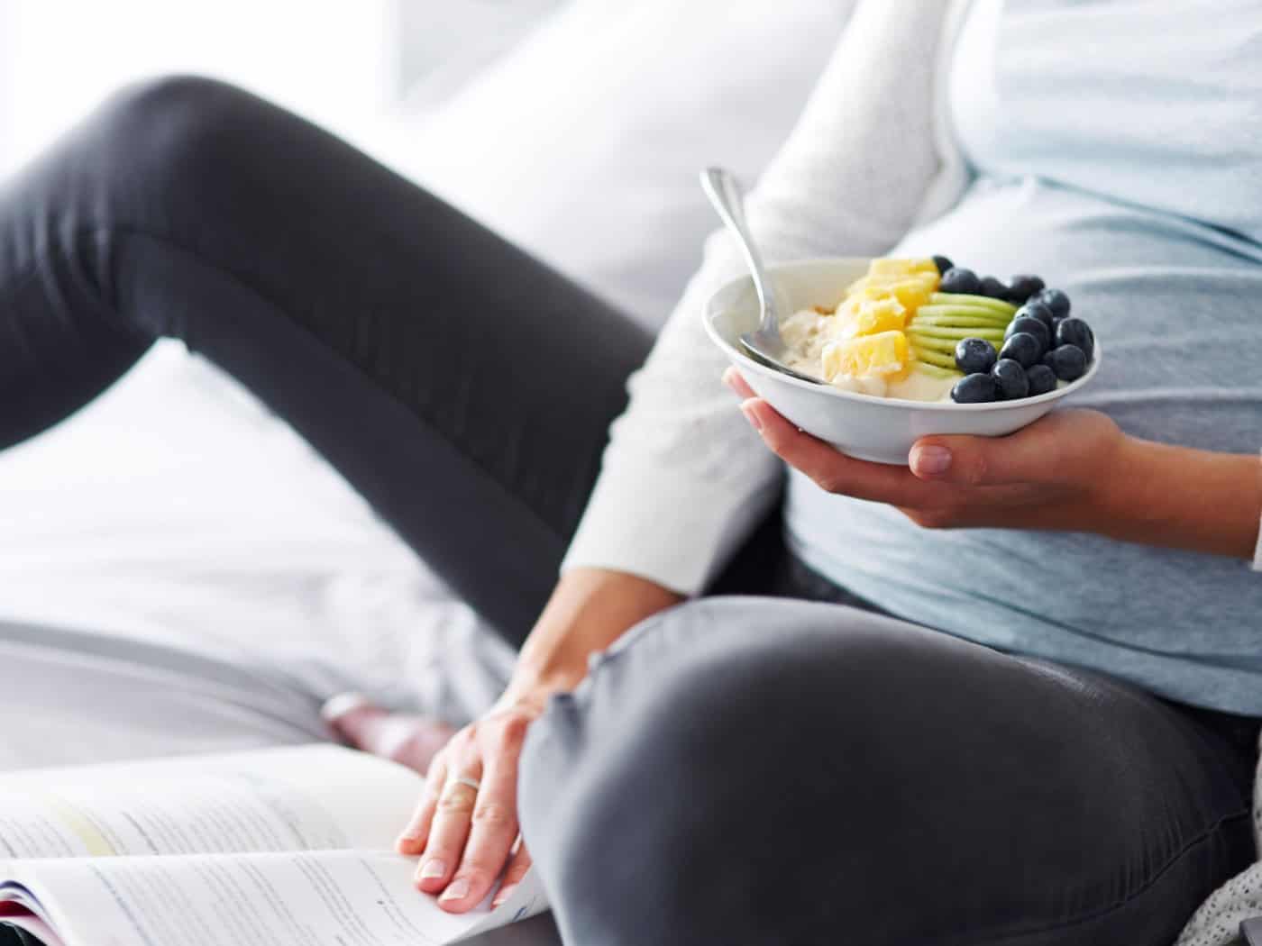 Nahaufnahme einer schwangeren Frau, die gemütlich im Bett sitzt und eine weiße Schale mit Müsli, Ananas-, Kiwi- und Blaubeerstücken in den Händen hält. Vor ihr liegt ein aufgeschlagenes Buch auf der Decke.