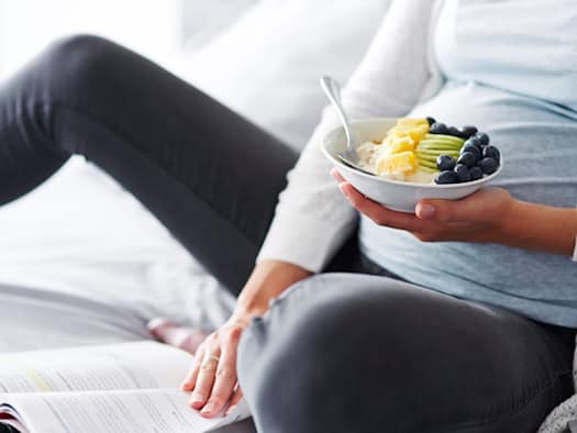 Nahaufnahme einer schwangeren Frau, die gemütlich im Bett sitzt und eine weiße Schale mit Müsli, Ananas-, Kiwi- und Blaubeerstücken in den Händen hält. Vor ihr liegt ein aufgeschlagenes Buch auf der Decke.
