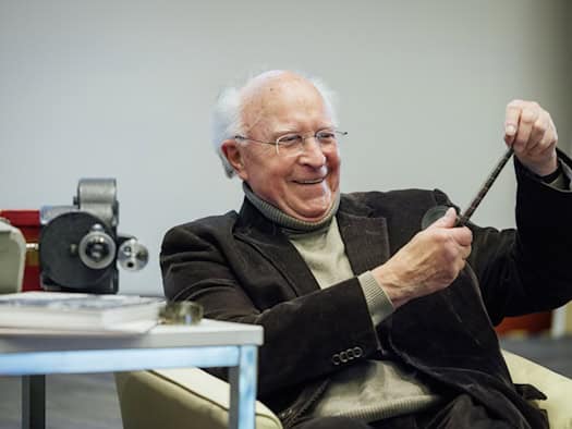 Ein älterer Mann mit Brille und Cord-Sakko lächelt, während er einen schmalen Filmstreifen mit beiden Händen gegen das Licht hält. Im Hintergrund ist eine historische Filmkamera auf einem Tisch erkennbar.
