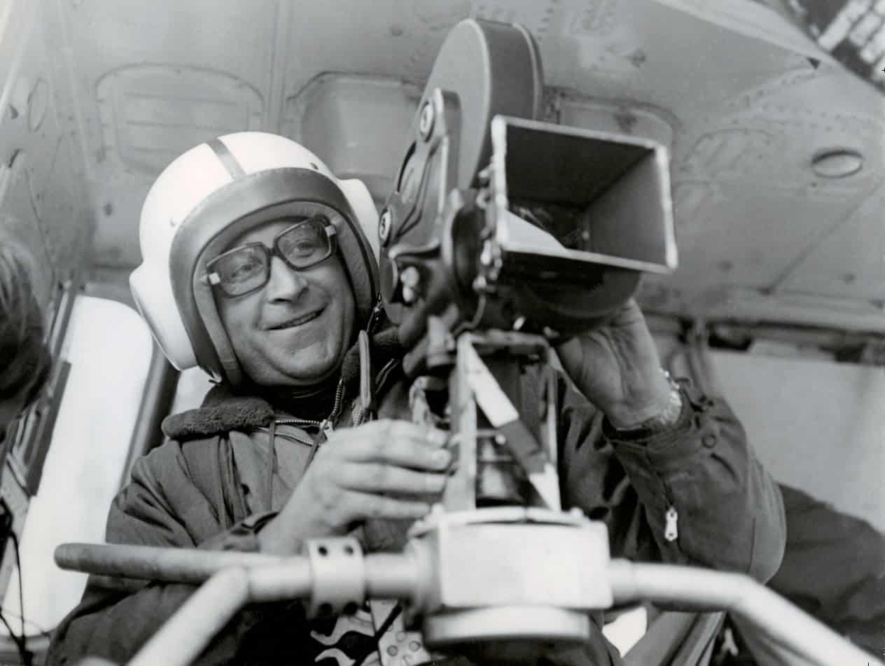 Schwarz-Weiß-Fotografie aus dem Jahr 1970: Ernst Hirsch trägt einen hellen Schutzhelm mit Brille und bedient lächelnd eine professionelle Filmkamera, die auf einem Stativ im Inneren eines Hubschraubers montiert ist.