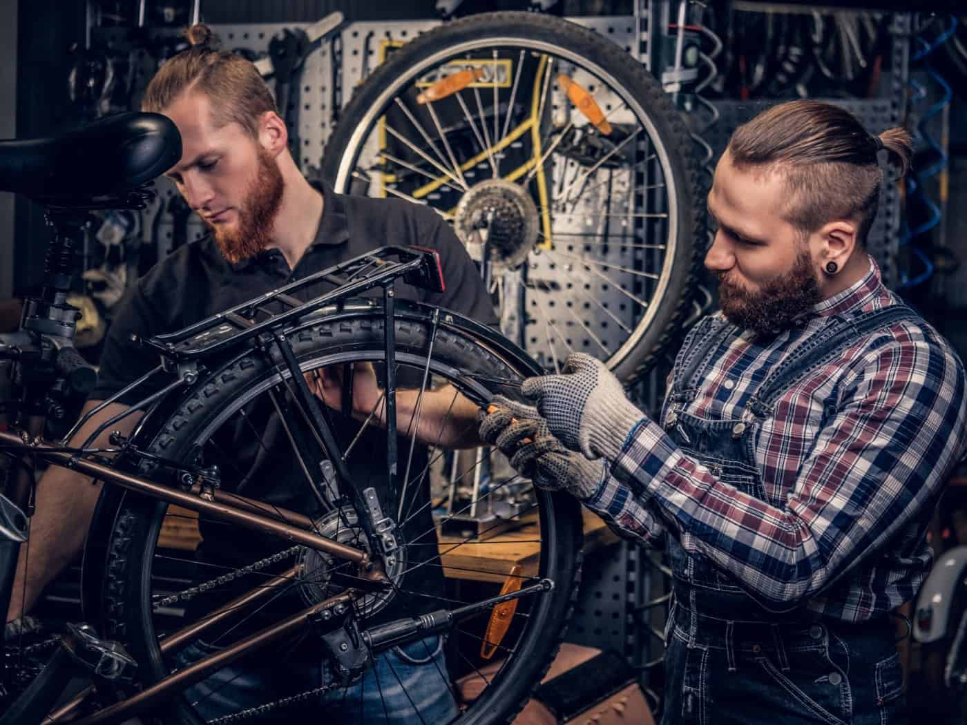 Zwei Fahrradmechaniker mit Bärten und hochgebundenen Haaren arbeiten konzentriert in einer Werkstatt an einem aufgebockten Tourenrad. Im Hintergrund hängen Werkzeuge und Ersatzteile an einer Lochwand.