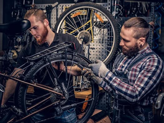 Zwei Fahrradmechaniker mit Bärten und hochgebundenen Haaren arbeiten konzentriert in einer Werkstatt an einem aufgebockten Tourenrad. Im Hintergrund hängen Werkzeuge und Ersatzteile an einer Lochwand.