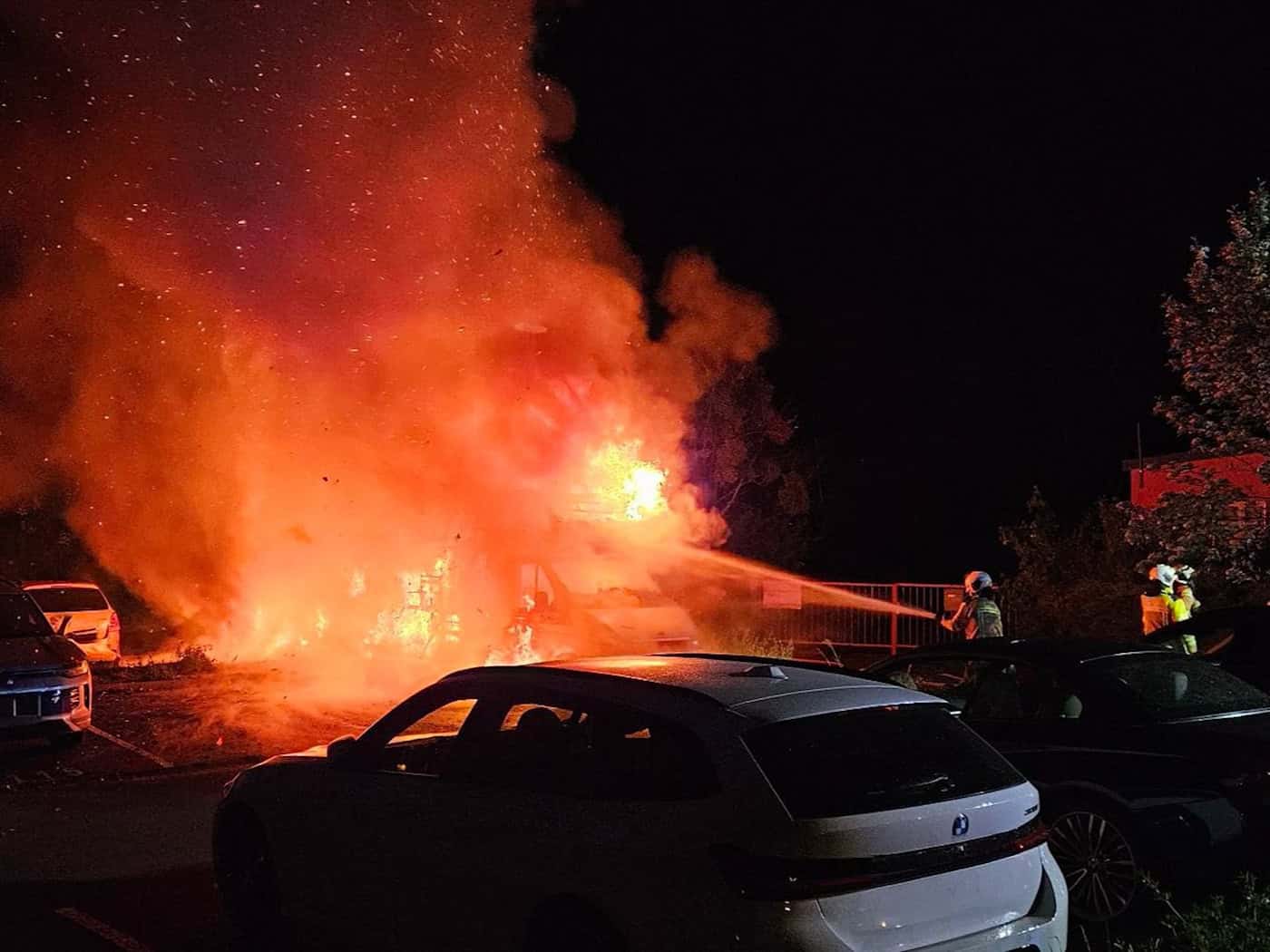 Nächtlicher Feuerwehreinsatz auf einem Parkplatz in Dresden. Ein Wohnmobil steht in hellem Vollbrand, die Flammen schlagen meterhoch empor und erzeugen starken Funkenflug. Ein Feuerwehrmann in Schutzkleidung bekämpft das Feuer mit einem Wasserstrahl aus einem Strahlrohr. Im Vordergrund sind die Silh