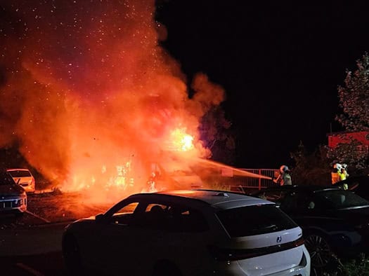 Nächtlicher Feuerwehreinsatz auf einem Parkplatz in Dresden. Ein Wohnmobil steht in hellem Vollbrand, die Flammen schlagen meterhoch empor und erzeugen starken Funkenflug. Ein Feuerwehrmann in Schutzkleidung bekämpft das Feuer mit einem Wasserstrahl aus einem Strahlrohr. Im Vordergrund sind die Silh